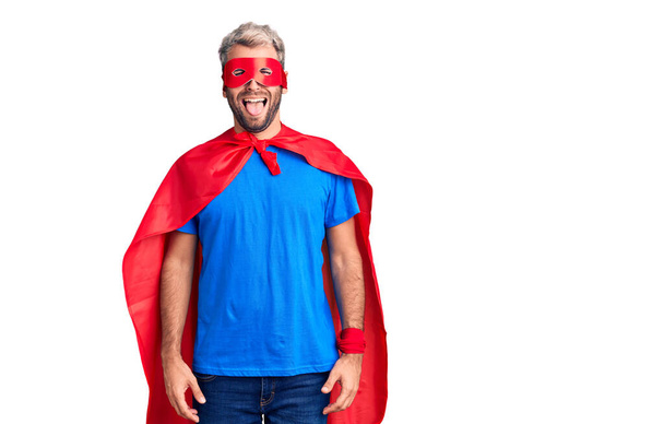 超ヒーローのカスタムを身に着けている若いブロンドの男は面白い表情で幸せ舌を突き出す。感情の概念.  - 写真・画像