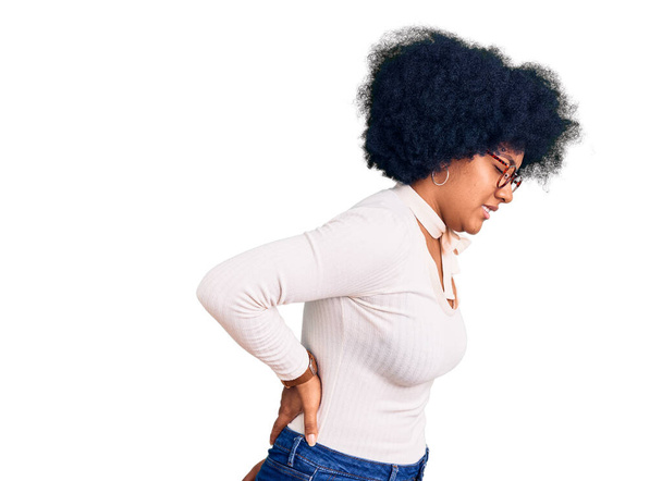Νεαρή Αφρικάνα Αμερικανίδα που φοράει καθημερινά ρούχα και γυαλιά και υποφέρει από πόνο στην πλάτη, αγγίζοντας την πλάτη με το χέρι, μυϊκό πόνο  - Φωτογραφία, εικόνα