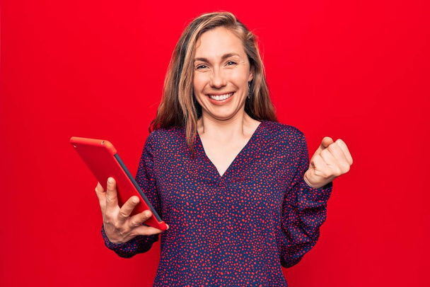 Fiatal szép szőke nő használ tabletta felett elszigetelt piros háttér sikoltozó büszke, ünneplő győzelem és a siker nagyon izgatott felemelt kar  - Fotó, kép