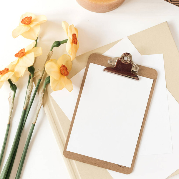 Blank paper sheet clipboard tablet pad with mock up copy space. Beautiful home office table desk workspace with fresh narcissus flowers, wooden casket, candle, albums. Flat lay, top view art concept - Valokuva, kuva