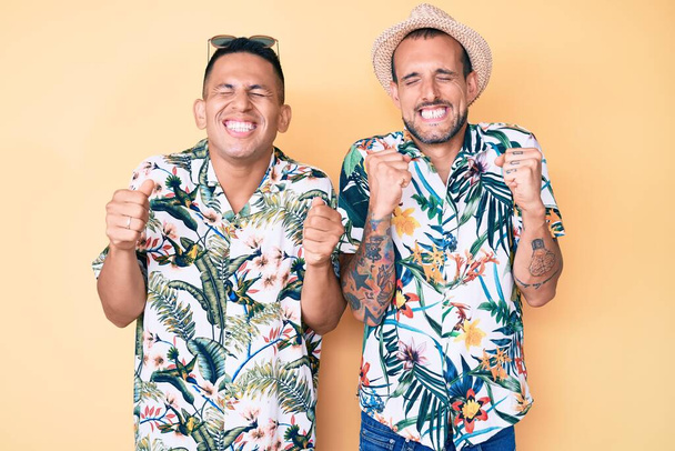 Das junge schwule Paar aus zwei Männern mit Sommermütze und hawaiianischem Hemd fiebert dem Erfolg entgegen, mit erhobenen Armen und geschlossenen Augen feiert es lächelnd den Sieg. Siegerkonzept.  - Foto, Bild