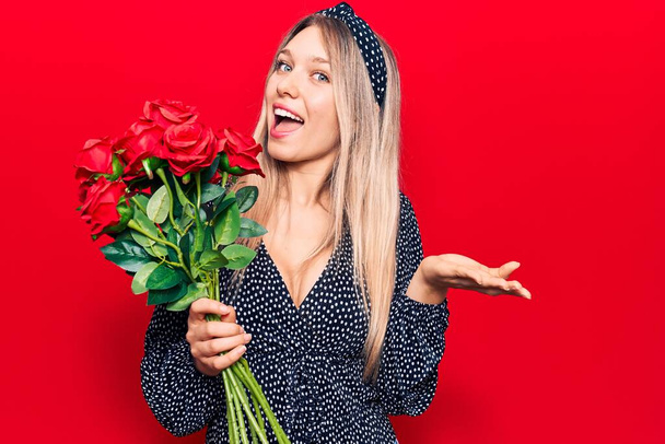Joven mujer rubia sosteniendo flores celebrando el logro con sonrisa feliz y expresión ganadora con la mano levantada  - Foto, Imagen