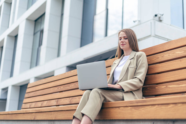 Beautiful blonde business woman in jacket working at laptop sit down on bench outside on a urban city street. Happy lady girl distance learning, education and online shopping. - Photo, Image