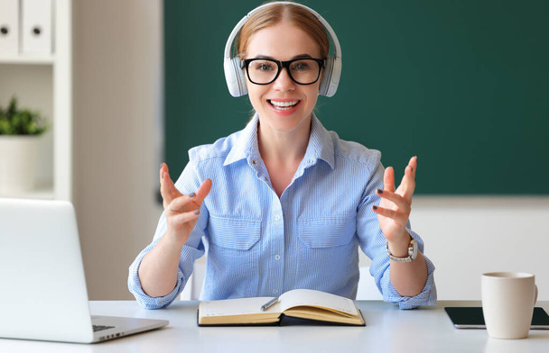 Mulher adulta feliz em óculos e fones de ouvido sorrindo e falando com a câmera enquanto sentada na mesa durante a aula on-line em sala de aula - Foto, Imagem