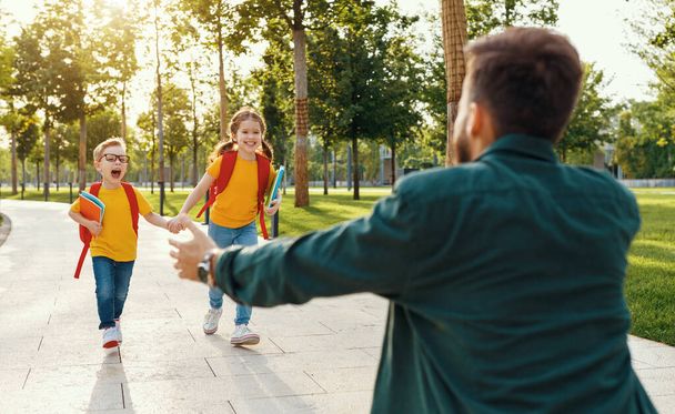 Чоловік зустрічає веселого хлопчика і дівчинку з блокнотами, що бігають на руках, зустрічаючи дітей після навчання в школі в парі
 - Фото, зображення