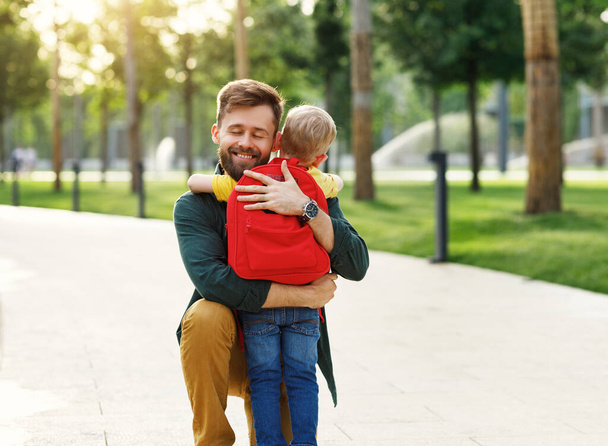 Vrolijke gelukkige vader glimlachend en omhelzend zoon schooljongen met rugzak voor school, terwijl knielen op pad in groene par - Foto, afbeelding