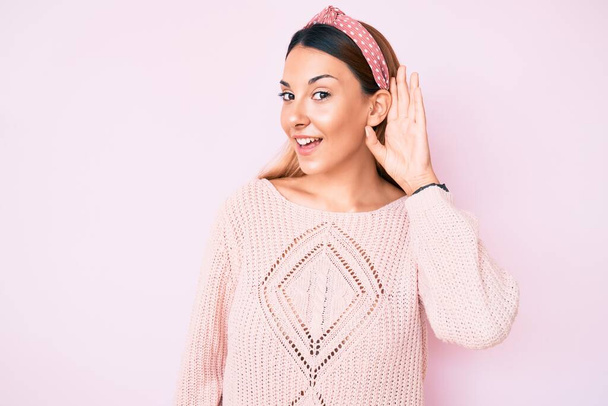 ピンクの背景にカジュアルな服を着た若いブルネットの女性が耳を傾け、噂やゴシップに耳を傾けながら手で笑っています。聴覚障害の概念.  - 写真・画像