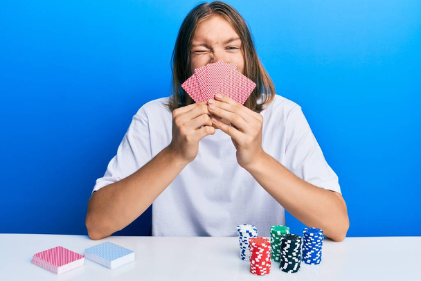 Uzun saçlı, kumarda poker oynayan, yüzünü kartlarla kaplayan yakışıklı beyaz adam kameraya seksi bir ifadeyle bakıyor, neşeli ve mutlu bir yüz..  - Fotoğraf, Görsel