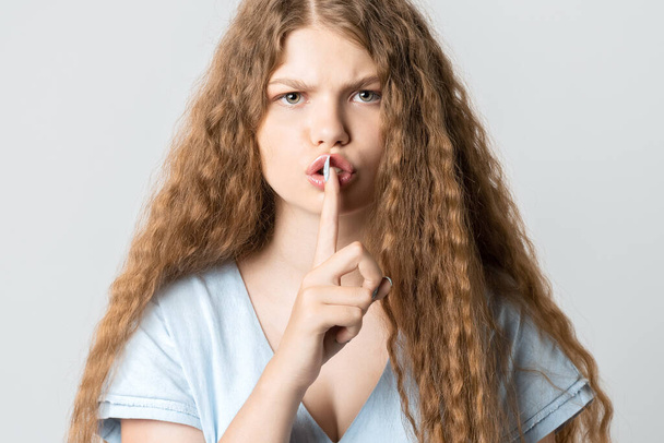 Emotional European young woman with curly long hair dressed in casual clothes, keeping index finger at her lips, saying Shh, Keep silent, It's secret - Fotografie, Obrázek