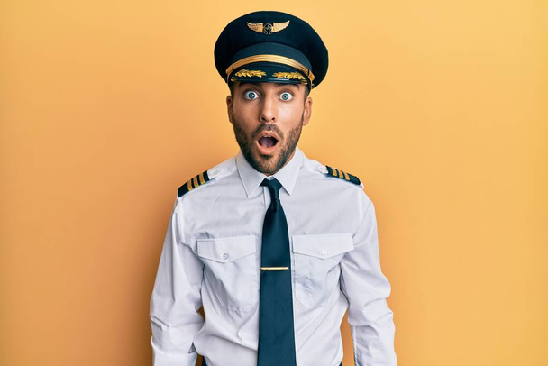Bel homme hispanique portant l'uniforme de pilote d'avion effrayé et choqué par la surprise et l'expression étonnante, la peur et le visage excité.  - Photo, image