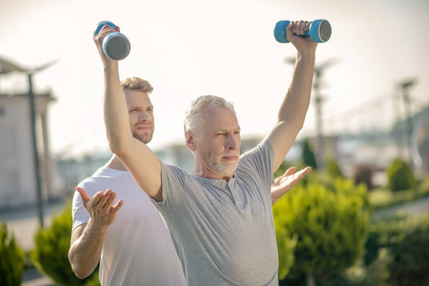 Grey-haired man raising hands with dumbbells, bearded instructor helping him - Foto, Bild