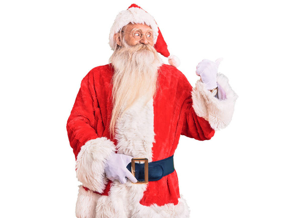 Velho homem sênior com cabelos grisalhos e barba longa vestindo traje tradicional de Papai Noel sorrindo com o rosto feliz olhando e apontando para o lado com o polegar para cima.  - Foto, Imagem