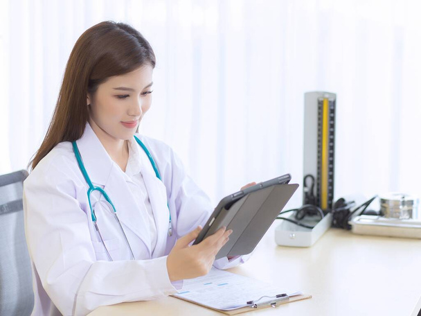 Ázsiai női orvos munkajelentése a betegéről tablettán. Van egy írótábla és egy vérnyomásmérő az asztalon a kórházban.. - Fotó, kép