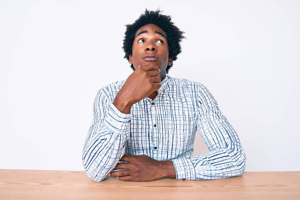 Jóképű afro-amerikai férfi afro hajjal, alkalmi ruhában, az asztalon ülve, komoly arccal, a kérdésre gondolva, kézzel az állon, átgondolva a zavaros ötletet.  - Fotó, kép