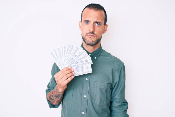 Young handsome man with tattoo holding dollars thinking attitude and sober expression looking self confident  - Photo, image