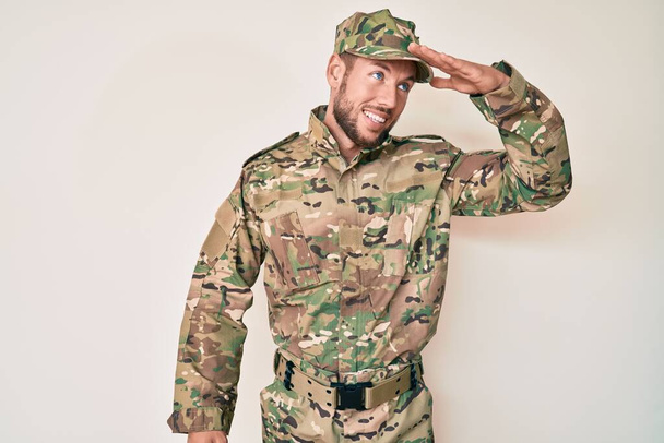 Young caucasian man wearing camouflage army uniform very happy and smiling looking far away with hand over head. searching concept.  - Фото, изображение