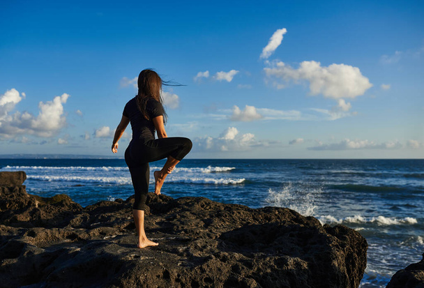 Το κορίτσι ασχολείται με τη γιόγκα και στέκεται σε ένα asana στο ένα πόδι με τα χέρια της ψηλά στις πέτρες με θέα τη θάλασσα και τον γαλάζιο ουρανό - Φωτογραφία, εικόνα