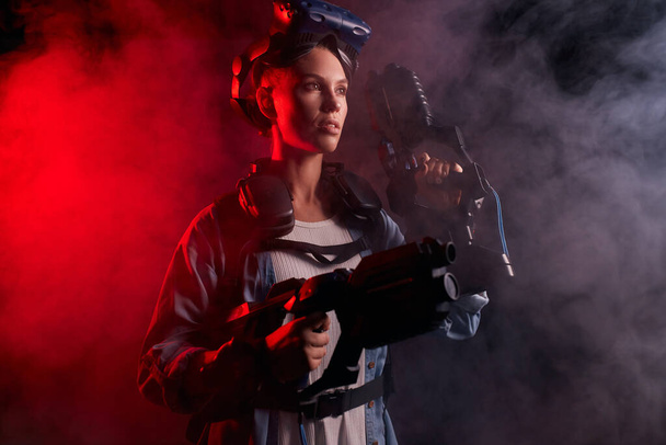 Young female playing game using virtual reality headset and guns in the dark smoky room - Foto, Imagem