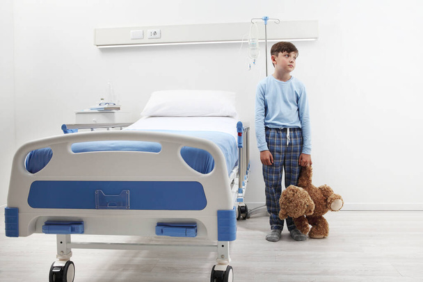 Gyerek a kórházi szobában áll az ágy mellett egy plüssmackóval, egy fehér falon elszigetelt pizsamában, fénymásolási hellyel.  - Fotó, kép