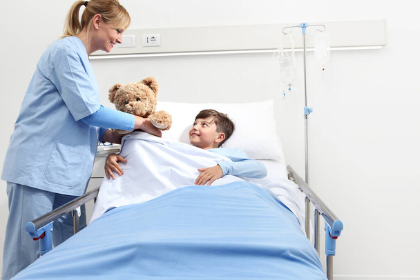 Uśmiechnięta pielęgniarka daje misia szczęśliwemu dziecku leżącemu w łóżku w pokoju szpitalnym na białym tle z przestrzenią do kopiowania - Zdjęcie, obraz