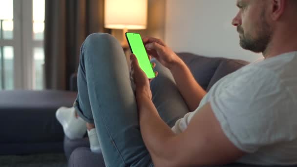 Homme à la maison couché sur un canapé et utilisant un smartphone avec écran vert maquette en mode vertical. Il navigue sur Internet, regarde du contenu, des vidéos, des blogs. Point de vue. - Séquence, vidéo