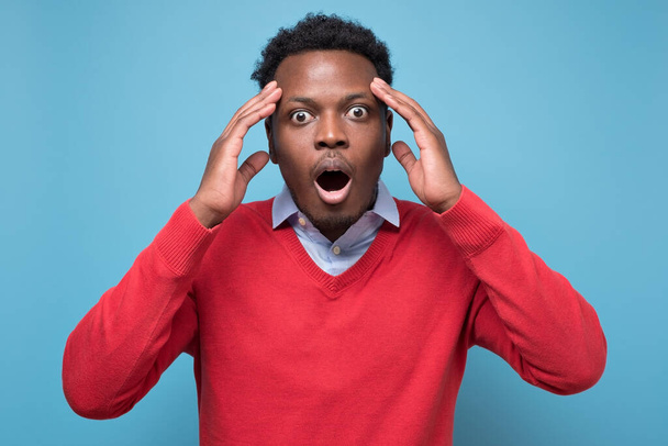 Взволнованный африканский американец в красном свитере в панике, смотрит с подслушивающими глазами, шокирован, услышав неожиданные новости. Великое удивление или неверие - Фото, изображение