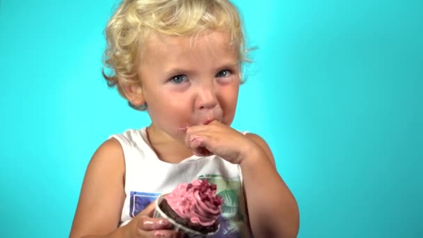 Un simpatico bambino maschio di 1 anno con gli occhi azzurri e i capelli biondi lecca con il dito un delizioso muffin al lampone. Mangia con le mani su uno sfondo blu. - Filmati, video