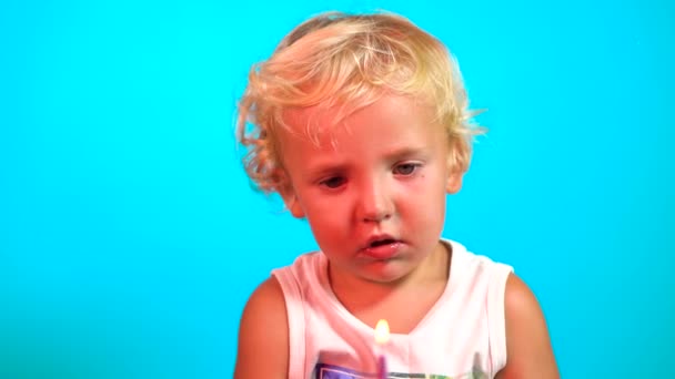 Bellissimo bambino maschio di 1 anno con gli occhi azzurri e i capelli biondi soffia una candela divertente su un muffin al lampone. All'interno sfondo blu. - Filmati, video