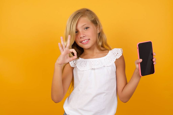 симпатичная девушка, держащая в руках смартфон и показывающая все хорошо на желтом фоне  - Фото, изображение