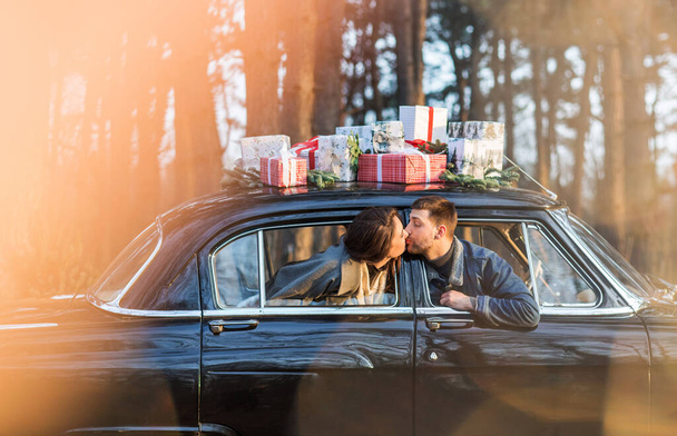 Loving man and woman sticking out of retro car windows and kissing with Christmas gifts on roof in park - Photo, Image