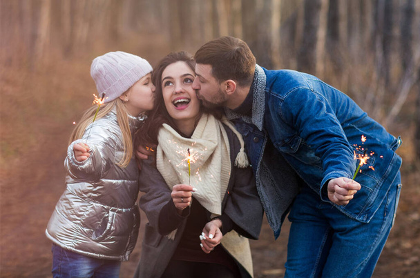 Joyeux jeune famille avec fille vêtue de vêtements chauds avec des étincelles dans les mains embrassant et regardant la caméra tout en se tenant debout sur fond de forêt - Photo, image