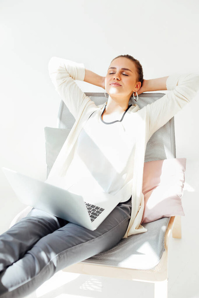 Jeune femme avec les mains derrière la tête assise sur la chaise et relaxant avec les yeux fermés après avoir utilisé un ordinateur portable dans la pièce lumineuse à la maison - Photo, image