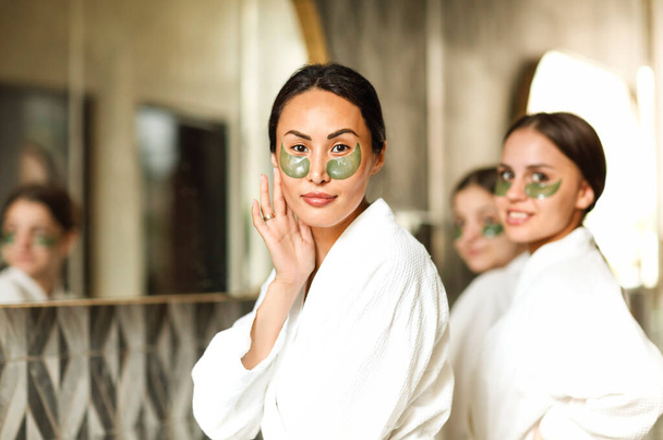 Towarzystwo uśmiechniętych kobiet w szlafrokach i opaskach na oczy stojących obok lustra w łazience i patrzących w kamerę podczas codziennej pielęgnacji skóry - Zdjęcie, obraz