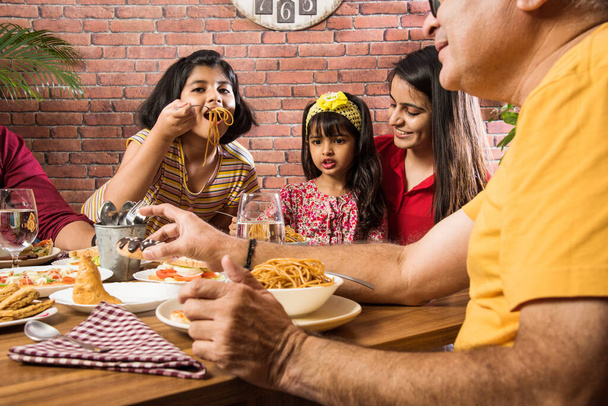 インドの多世代家庭やレストランのダイニングテーブルで食べ物を食べる家族。南アジアの祖父、母、父と2人の娘が一緒に食事をしている - 写真・画像