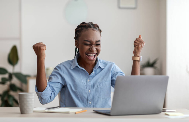 Euforisch zwart vrouwelijke werknemer viert succes met laptop op het werk in Office - Foto, afbeelding