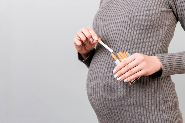 Großaufnahme einer schwangeren Frau, die vor grauem Hintergrund eine Zigarette aus einer Packung nimmt. Ungesunder Lebensstil während der Schwangerschaft. Rauchersucht. - Foto, Bild