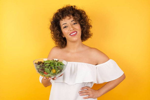 Studio shot of cheerful woman holding a salad dressed casual keeps hand on hip, smiles broadly, anticipates for surprise prepared by friends on her birthday, isolated over yellow  background. - 写真・画像