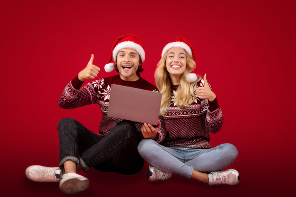Compras de Navidad en línea. Pareja joven en sombreros de Santa con ordenador portátil que muestra los pulgares hacia arriba sobre el fondo rojo - Foto, imagen