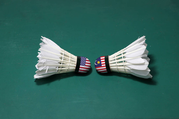 Duas vaivéns usadas no chão verde da corte de Badminton com ambas as cabeças uma na outra. Uma cabeça pintada com bandeira dos EUA e uma cabeça pintada com a bandeira da Malásia, conceito de competição de badminton. - Foto, Imagem