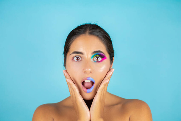 Beauty portrait of female model with vivid makeup on one half of the face and clean face on the other part on blue background and blue lips. - Photo, Image