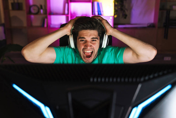 Image of unhappy young man in headphones grabbing his head while playing video game on computer at home - Фото, изображение