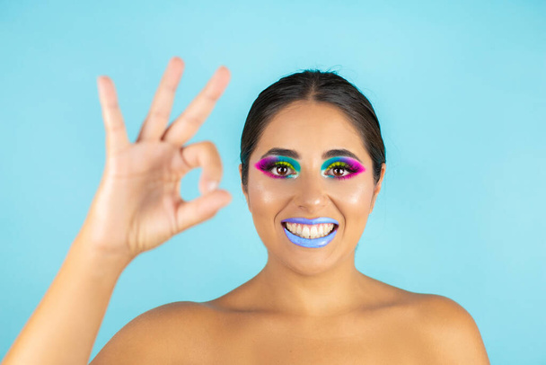 Schoonheidsportret van vrouwelijk model met levendige make-up op blauwe achtergrond en blauwe lippen. Een goed gebaar maken - Foto, afbeelding
