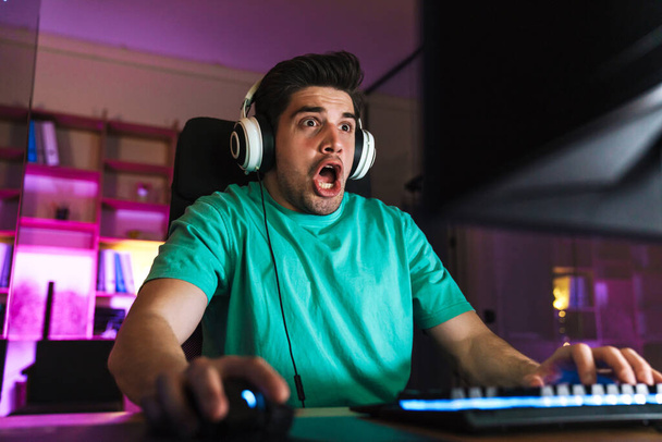 Image of shocked young man in headphones screaming while playing video game on computer at home - Foto, Imagen