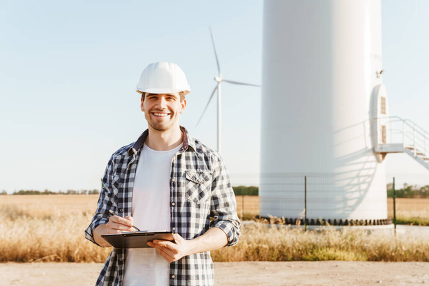A smiling electrical engineer standing on a field with windmills, taking notes - 写真・画像