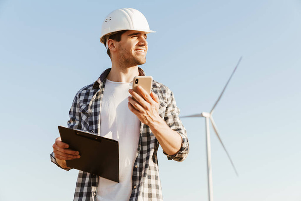 Ein lächelnder männlicher Ingenieur mit dem Handy, während er auf der Windkraftanlage steht - Foto, Bild