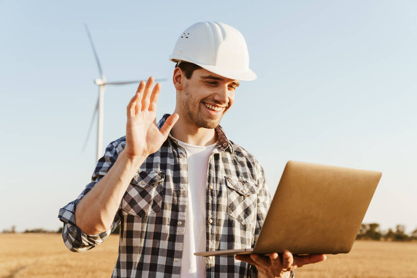 An electrical engineer standing on a field with windmills, using laptop computer, videocall, waving hand - Foto, Imagen