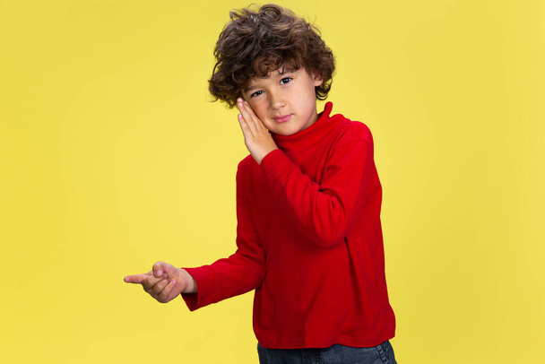 Pretty young curly boy in red wear on yellow studio background. Childhood, expression, fun. - Zdjęcie, obraz