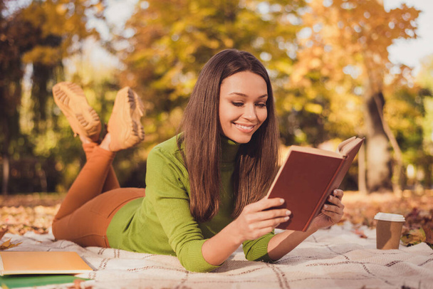 Photo of pretty charming dreamy lady autumn park leaves cover ground lay blanket among trees smiling reading fiction paper book drink coffee wear green turtleneck pants sneakers outdoors - Foto, Imagem