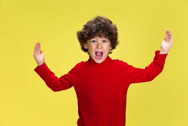 Pretty young curly boy in red wear on yellow studio background. Childhood, expression, fun. - Foto, imagen