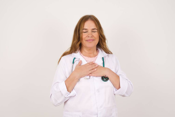 Faithful mature caucasian doctor woman wearing medical uniform closes eyes and keeps hands on chest near heart, shows her kindness or favor, expresses sincere emotions, being kind-hearted and honest. Body language and real feelings concept. - Photo, Image